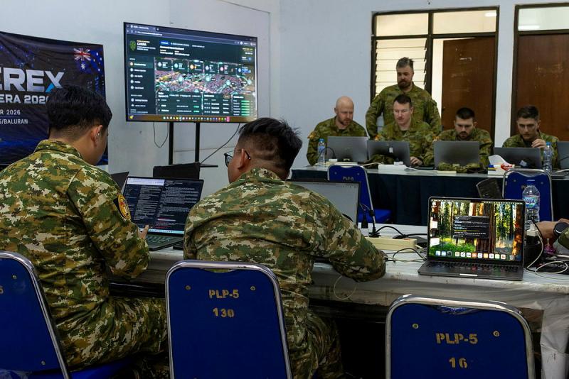Two small groups of military personnel sit behind desks on either side of a room, all working on laptops.