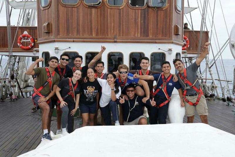 People wearing safety harnesses stand on the deck of a tall ship, smiling and giving thumbs up.