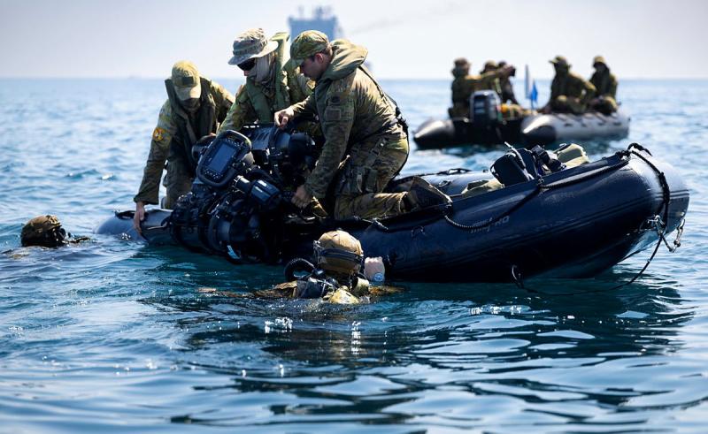 Soldiers lower diving gear into the water while onboard an inflatable raft. Divers wait in the ocean water.