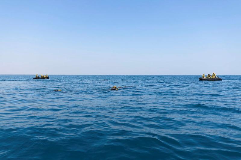 Two small boats float in the ocean with military personnel on board and divers in the water.