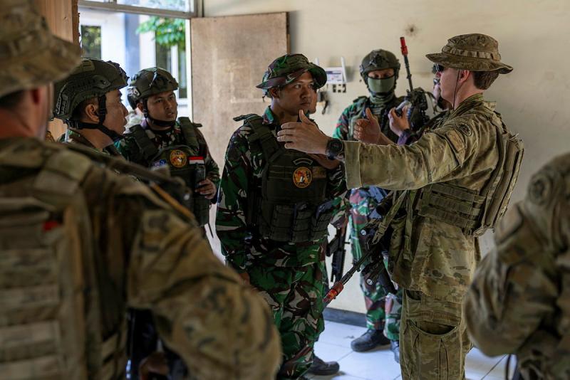 A group of military personnel standing in a building listening to another military man instructing.