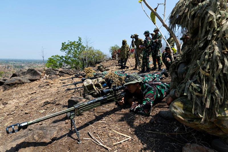 A man in uniform lies on the ground looking through the sight of a rifle with a very long barrel while others watch from behind.