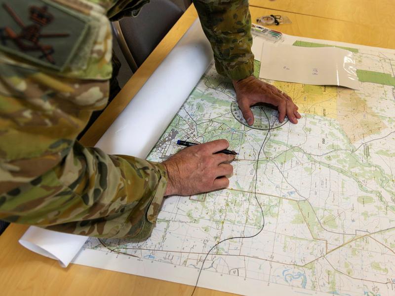 A person wearing Army uniform holds a protractor and pen to a map on a table.