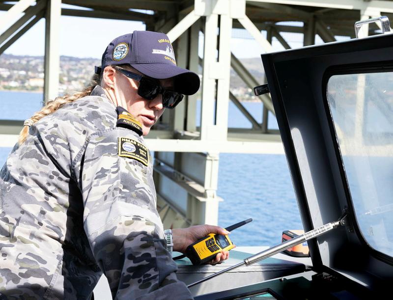 A woman in Navy uniform looks over her shoulder while holding a handheld radio.