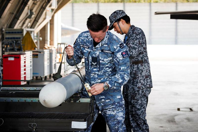 Two military men put a strap around a narrow cylindrical pole.