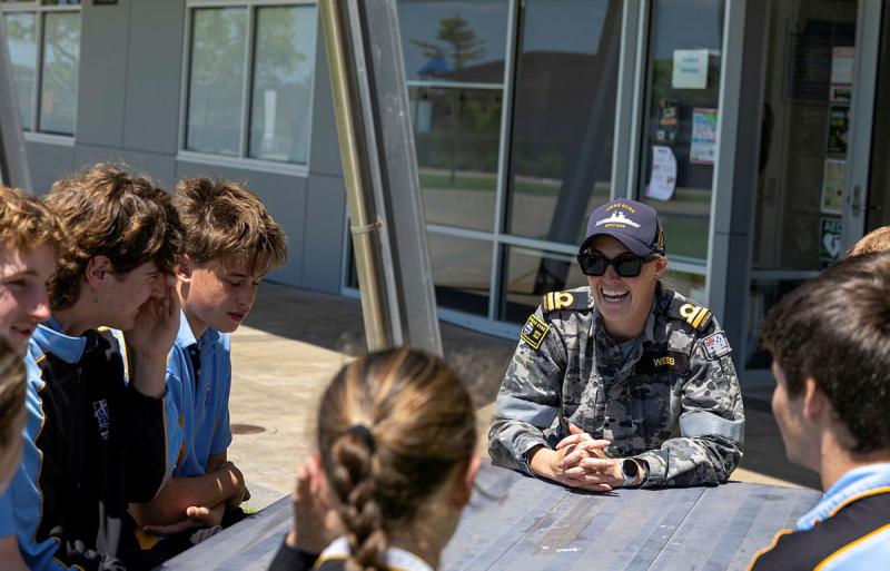 A person in a Navy uniform smailes and talks with a small group of children.