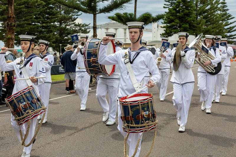 Navy personnel in whit uniforms play musical instruments as the march along a street.