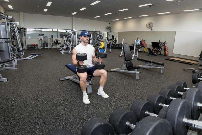 A man holds two dumbbells on his knees while in a gym area.
