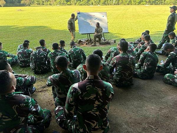 People in uniform sit on the ground outdoors while looking at two people standing at a whiteboard in front of them.