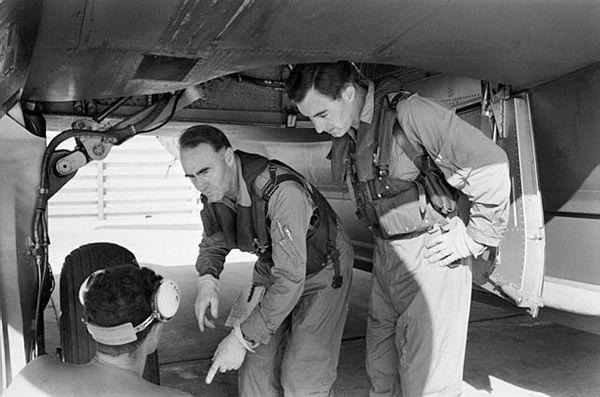 Two men crouch underneath an aircraft, one points at something towards the ground to another crouched man whose back is visible
