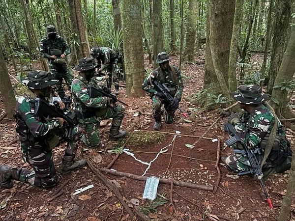 People in Indonesian Army camouflage uniform crouch in a jungle, looking at a makeshift grid map in the dirt.