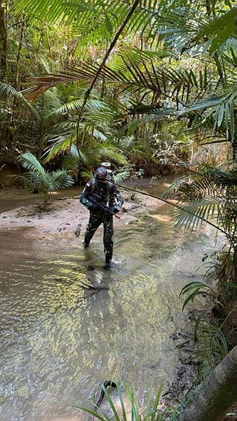 Two people in uniform cross a shallow stream in a jungle.