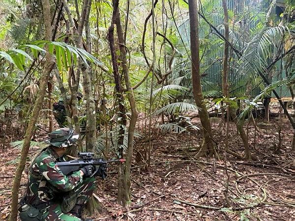 Two people in uniform move through dense jungle towards a building, weapons raised.