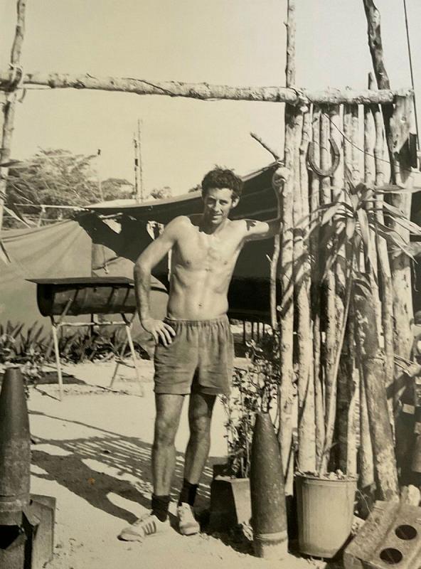 Bare-chested man in shorts stands under a frame made of large sticks with artillery shells on the ground nearby