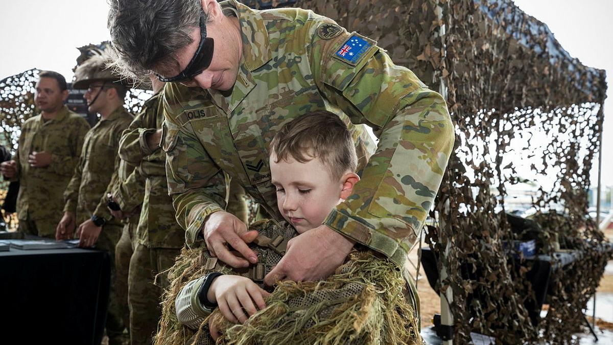 A man in Army uniform helps a young boy dress in camouflage. 