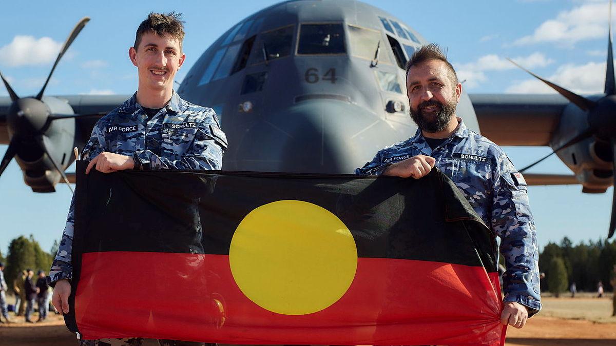 Two military men hold a First Nations flag in front of an aircraft.