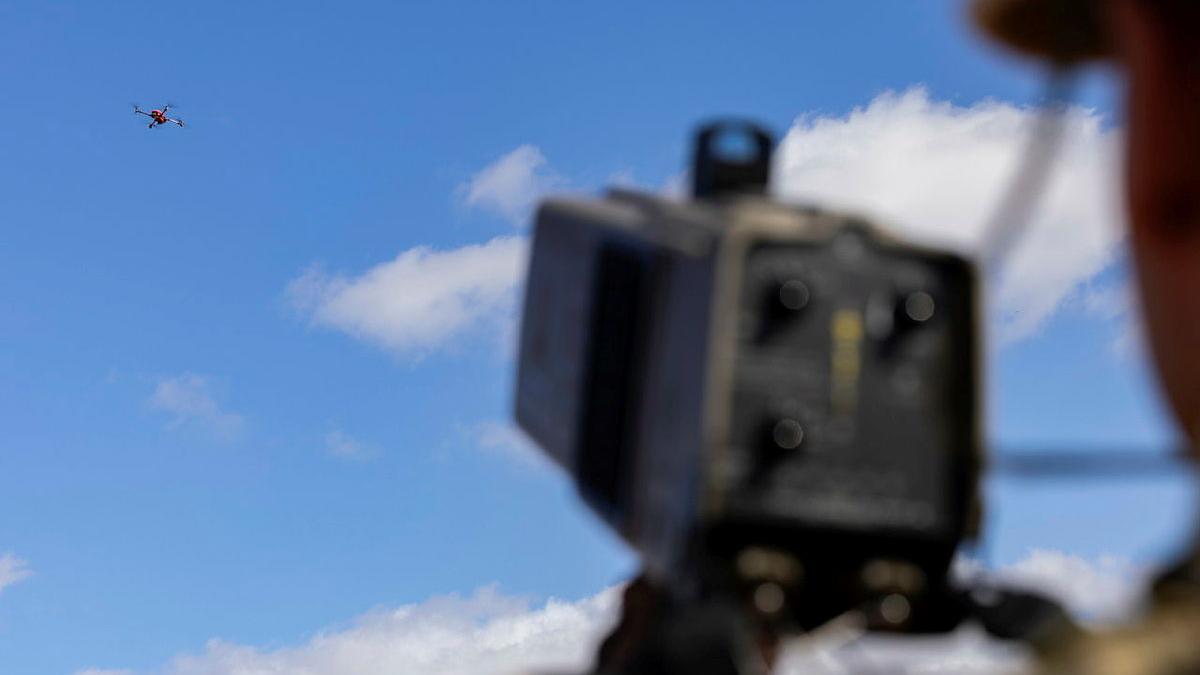 A soldier holds a scanner towards a drone in the sky.