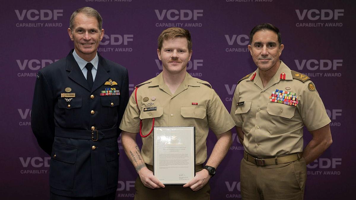 An Army private holds a framed certificate, with an Air Force chief and Army chief on either side of him.
