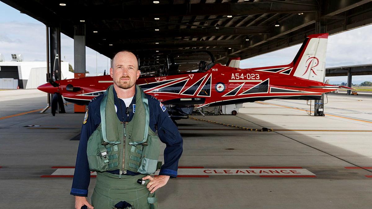 An Air Force pilot standing undercover on the tarmac in front of a red aircraft.