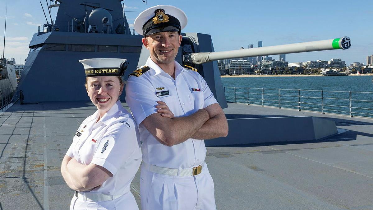 A male and female sailor stand back to back, smiling with their arms crossed, on the deck of a ship..