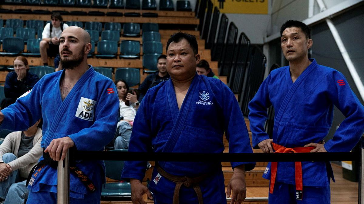 Three men in judo uniforms standing side by side watching a match. 