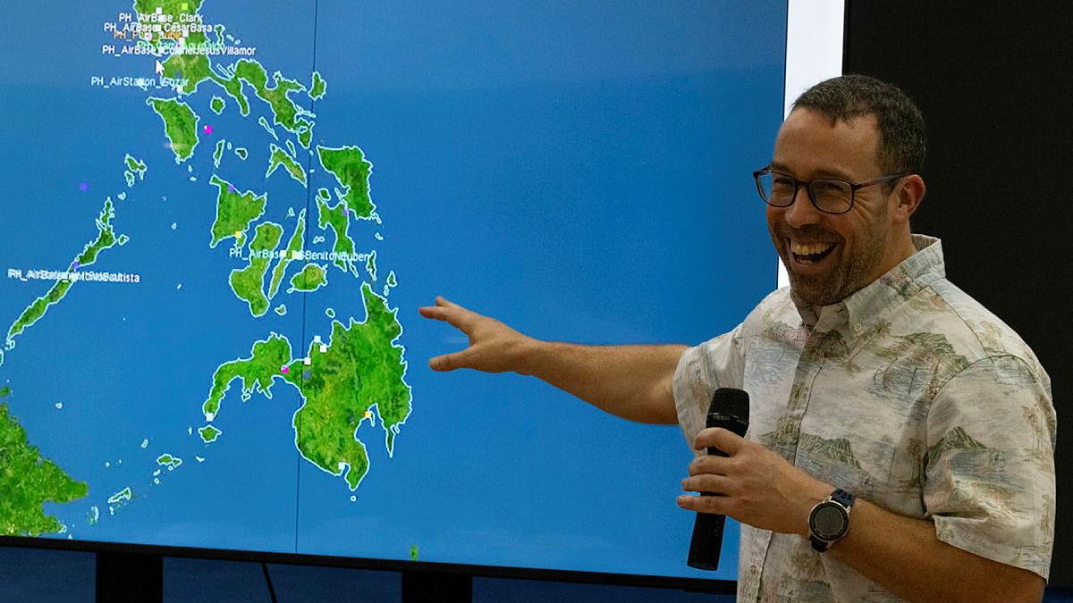 A man speaking with a microphone while pointing to a digital map displayed on the wall.