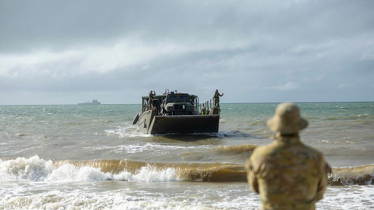 A solider stands on the beach as a landing craft approaches from the sea.