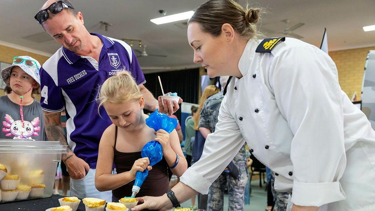 A sailor helps a child pipe icing onto a cupcake.