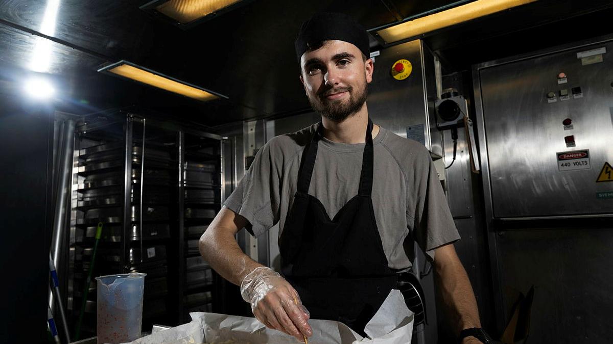 A Navy chef stands in an industrial kitchen and uses a skewer to for a pattern on the top of a cheesecake.