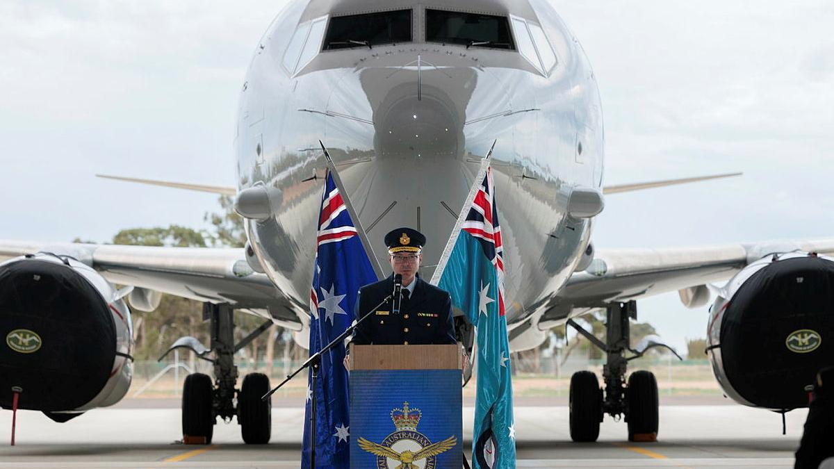 A man in military uniform speaks at a podium, with a large aircraft in the background.