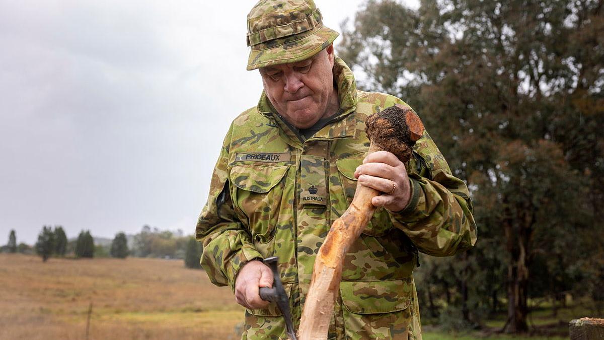 A man in camouflage uniform uses a small axe to shape a piece of wood.