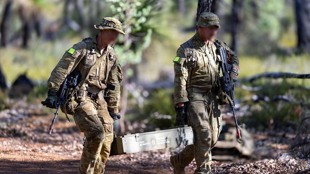Two people in military uniform carry a large metal box between them.