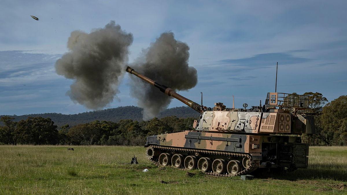 A tank in the bush fires ammunition, causing clouds of smoke on either side.