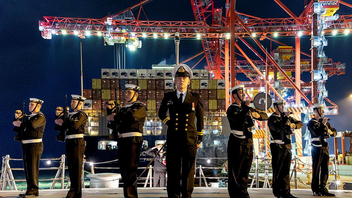 Navy personnel in black uniforms stand in formation in front of lit-up cranes and shipping containers..