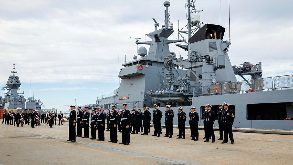 Sailors stand in formation on a concrete surface in front of a docked ship.