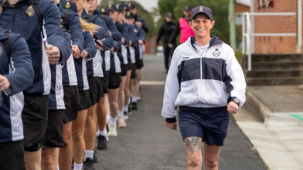 A woman in Navy exercise gear marches alongside a line of recruits in exercise uniform.