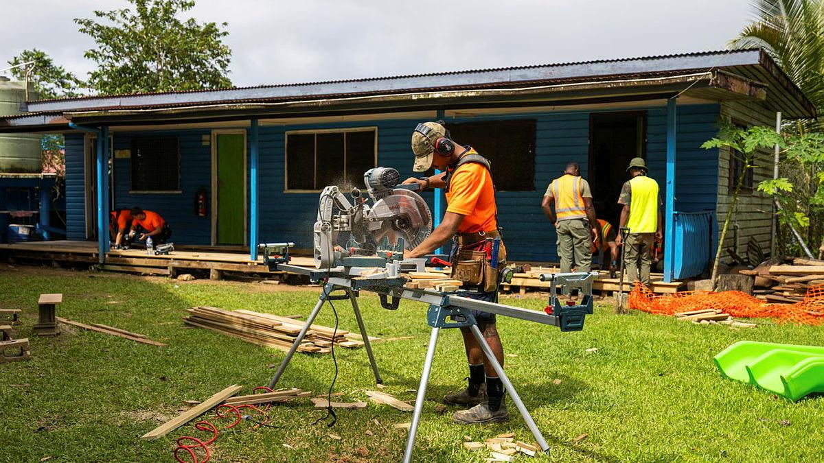 A man using a drop saw while outside a blue building.