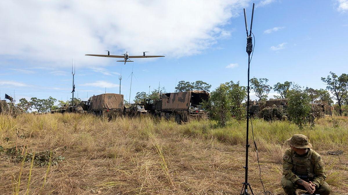 A small aircraft flies from behind towards a man in Army uniform crouching in front of a laptop computer.