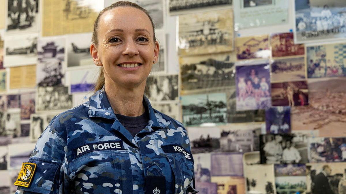 A female Air Force member stands, smiling, in front of a  display board of old photos.
