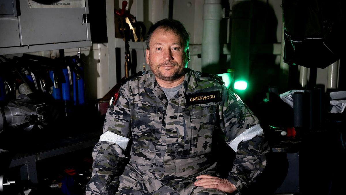 A Navy member in uniform sits with one hand on his hip in a room of equipment.