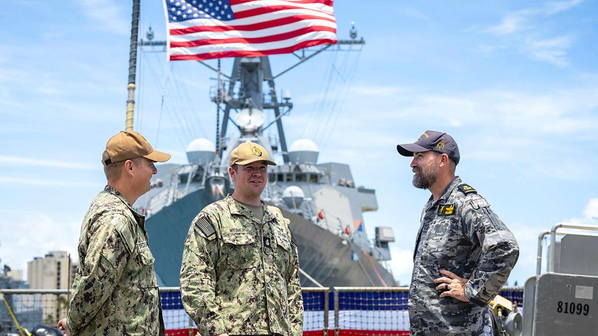 Three sailors speak to each other in front of a flag.