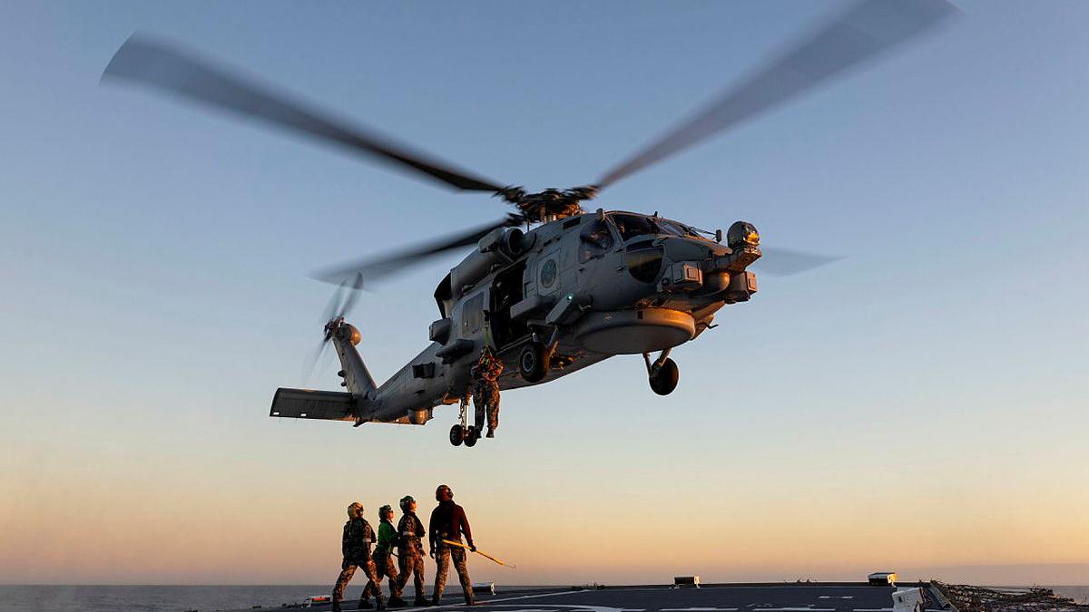 A helicopter hovers above a ship flight deck as a person hangs from a harness while four others stand below.