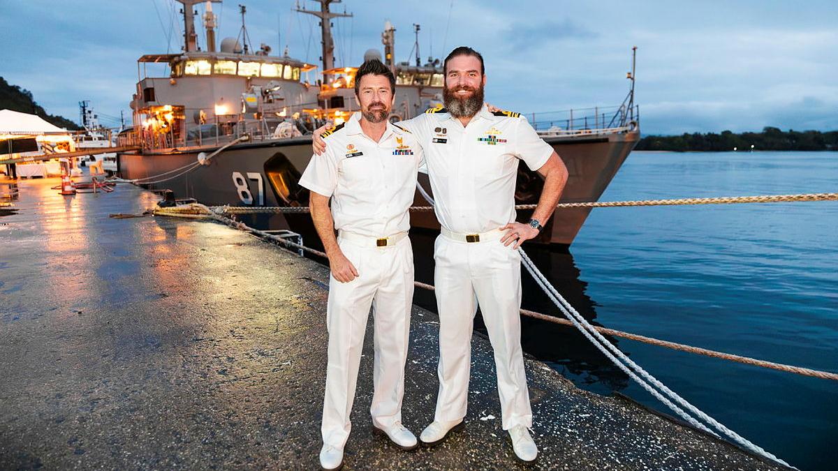 Two uniformed men stand on a wharf with their arms around each other's shoulders, with two ships behind them.