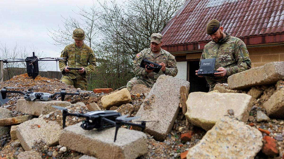 Army personnel standing in concrete rubble hold controls for drones.