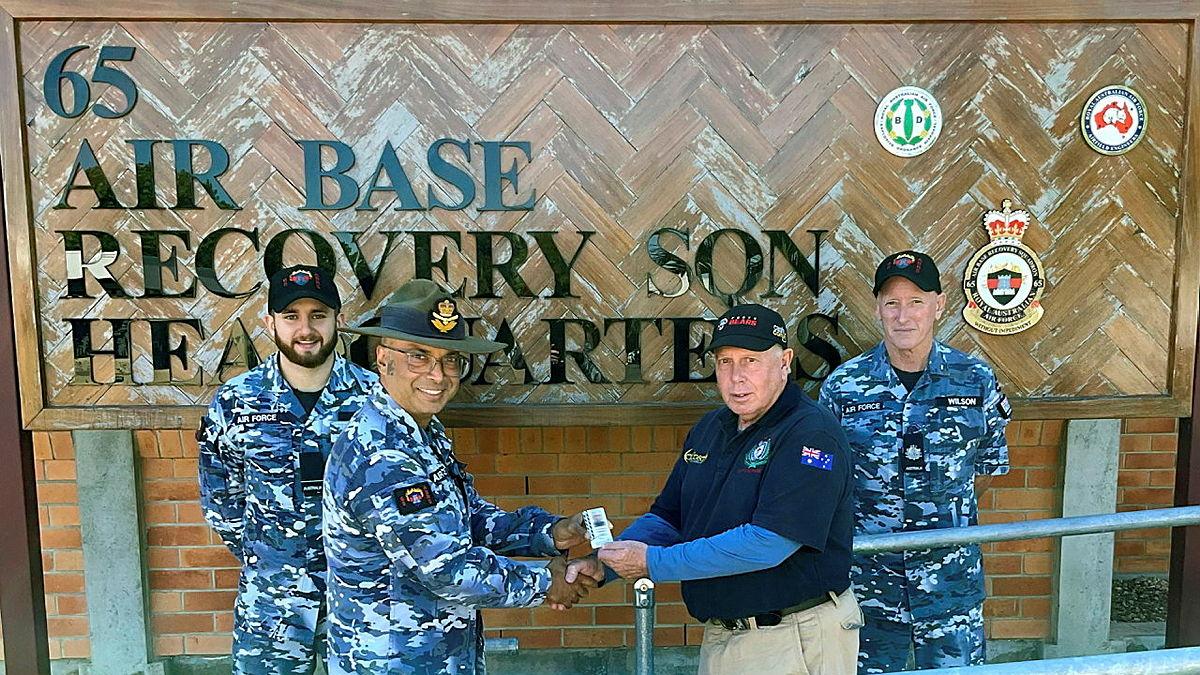 Military personnel give a small item to a civilian in front of a 65 Air base Recovery SQN Headquarters sign.