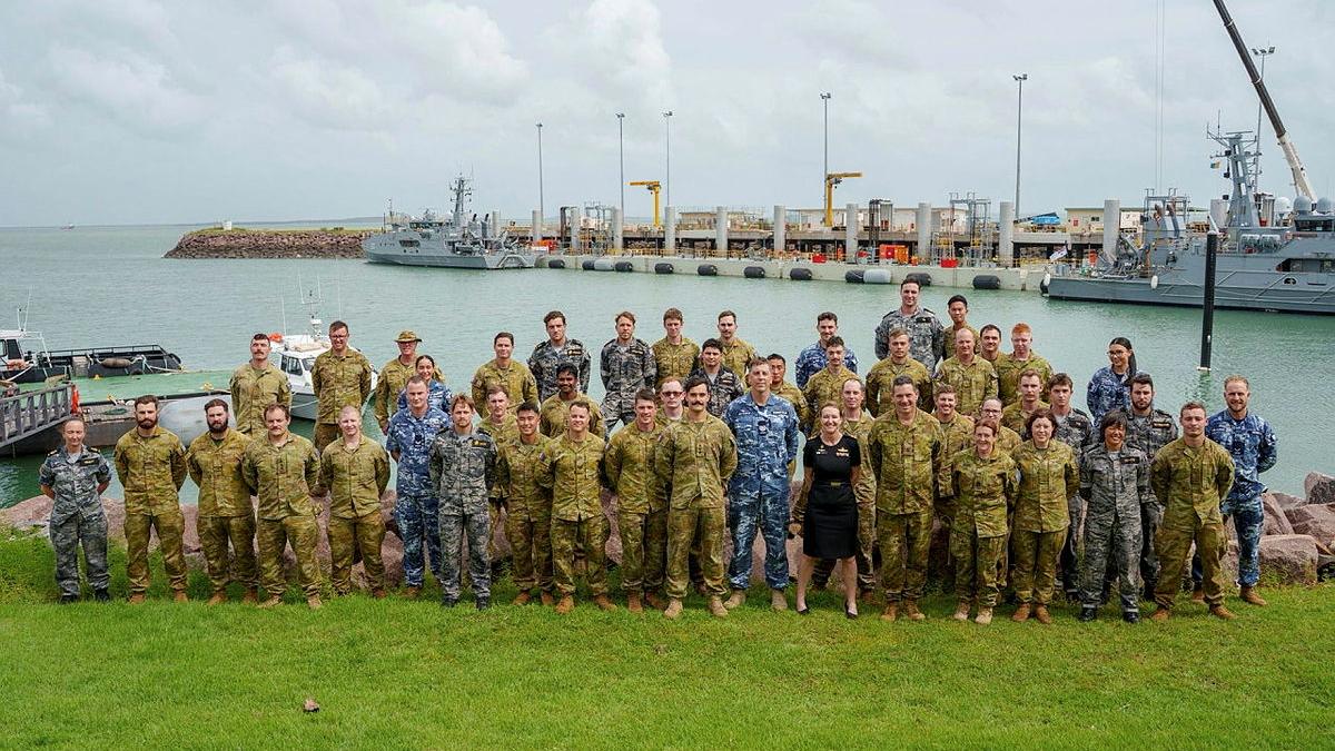 A large group of military personnel gather for a photo in front on water and ships.