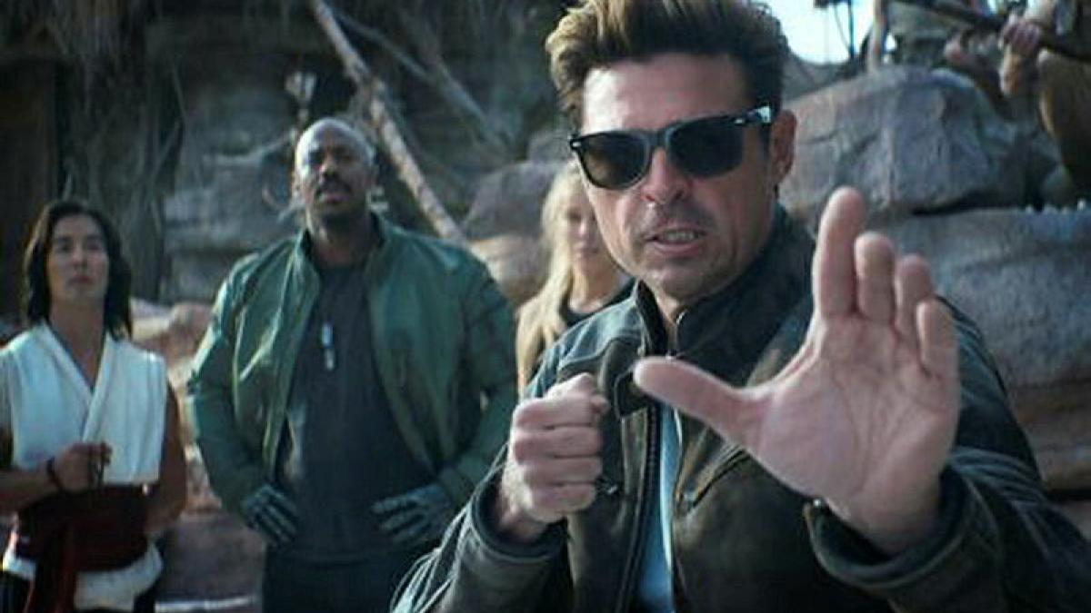 A man stands with one arm outstretched in front of him and the other in a fist my his side. A small group of people stand behind him.