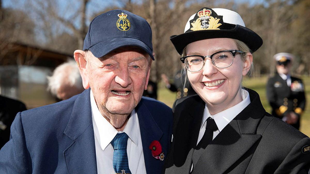 A military woman stands with an elderly man with a poppy pinned to his blazer.