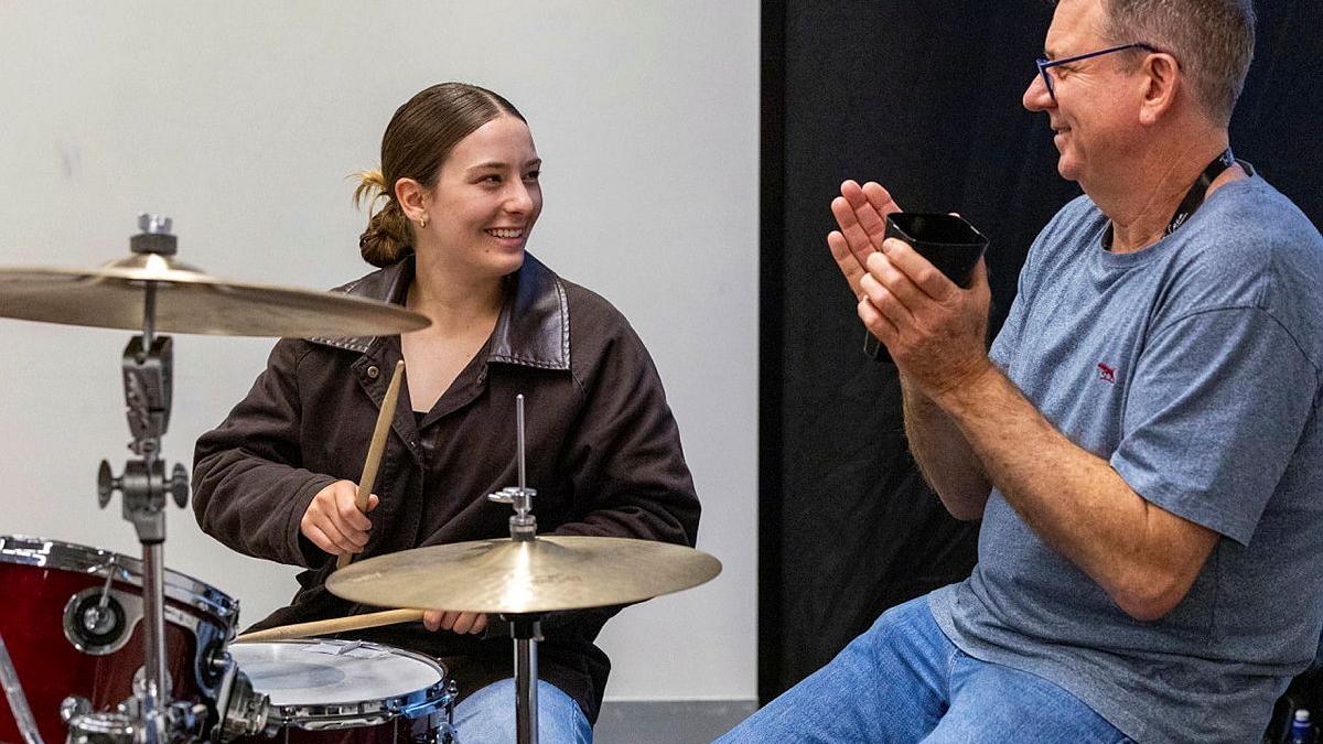 A seated man claps next to a young woman playing the drums.