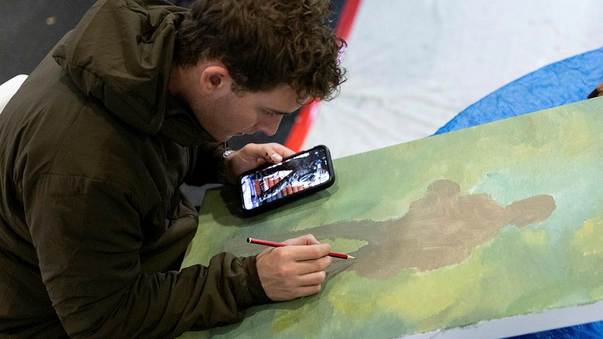 A man holds a phone in one hand while drawing on a painted canvas with a graphite pencil in the other.